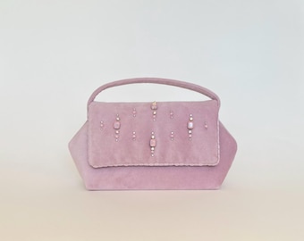 Op de afbeelding: Een lavendelkleurige handtas met een handvat aan de bovenkant en een klepsluiting. De tas is versierd met kleine, vierkante en ronde decoratieve elementen. De tas heeft een zachte, suède-achtige textuur en een gestructureerde vorm.