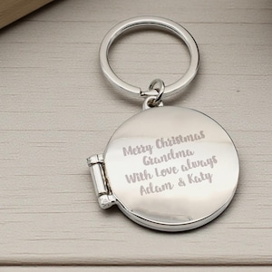 May include: A silver-colored, round locket keychain with a hinged opening. The locket has engraved text that reads "Merry Christmas Grandma With Love always Adam & Katy". The keychain includes a silver ring for attaching keys.