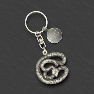 May include: Silver-coloured keychain with a coiled snake pendant. The snake has detailed scales and a small head. A round tag with the initials "MH" is attached. The keychain includes a split ring and a lobster clasp.