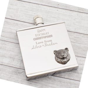 Puede incluir: Una petaca plateada con tapón de rosca. La petaca está grabada con "HAPPY BIRTHDAY CHRISTOPHER Love from Silvie & Graham". Una cabeza de tigre detallada está fijada en la esquina inferior derecha. La petaca es rectangular.