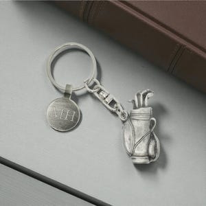 May include: Silver-coloured keychain with a golf bag charm containing golf clubs and a round tag engraved with "MH". The keychain is attached to a large ring. A brown leather-bound book is in the background.