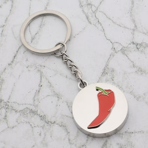Może przedstawiać: Srebrny brelok do kluczy z okrągłym zawieszką przedstawiającą czerwoną papryczkę chili z zielonym ogonkiem. Brelok ma srebrny pierścień i łańcuszek.
