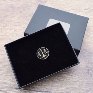 Puede incluir: Una caja de terciopelo negro con un pin de plata de la balanza de la justicia en su interior. El pin es un símbolo de la ley y la justicia.