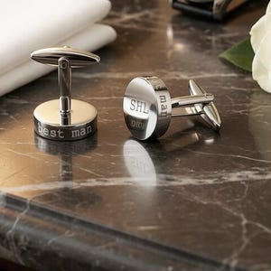 May include: Two silver cufflinks with a polished finish. One cufflink is engraved with "best man", the other with "SHL" and "man". The cufflinks are on a dark marble surface, with a white cloth and a flower in the background.