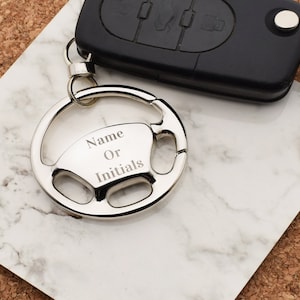 Könnte beinhalten: Silberner Autolenkrad-Schlüsselanhänger mit einem Ring und Platz für eine Gravur. Die Gravur lautet "Name oder Initialen".