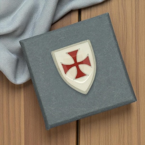 May include: A grey box featuring a white and red cross emblem. The emblem is a shield shape with a red cross on a white background. The box is on a wooden surface with a blue cloth.
