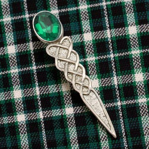 May include: A silver kilt pin with a Celtic knot design and a large, oval, emerald-green gemstone. The pin is displayed on a green, black, and white plaid fabric.