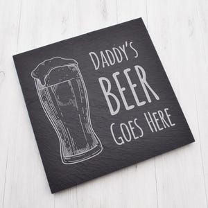 Puede incluir: Tabla de servir cuadrada de pizarra negra con el texto "DADDY'S BEER GOES HERE" en blanco. Un dibujo lineal detallado de un vaso de cerveza está en el lado izquierdo de la tabla. La tabla está sobre una superficie de madera blanca.