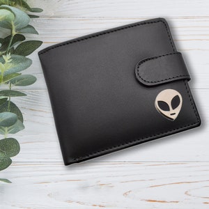 May include: Black leather wallet with a silver alien head design. The wallet has a fold-over flap with a snap closure.