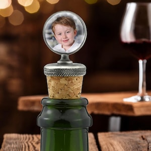 May include: A bottle stopper featuring a circular frame containing a photograph of a young child with light brown hair. The stopper has a textured silver-toned metal top and a natural light brown cork base, inserted into a dark green glass bottle.