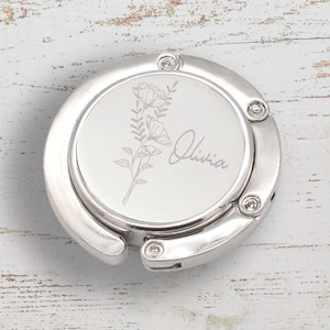 May include: A silver-coloured handbag hook featuring a floral design and the name "Olivia" engraved on the top. The circular hook has a folding mechanism. The background is a light-coloured, weathered wooden surface.