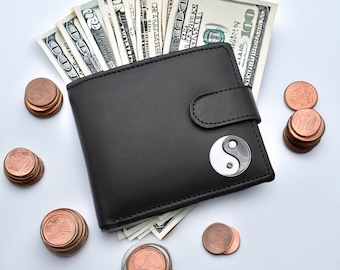 Op de afbeelding: Een zwarte leren portemonnee met een zilveren yin yang symbool aan de voorkant. De portemonnee is open en gevuld met Amerikaanse dollarbiljetten. Aan beide kanten van de portemonnee liggen stapels munten.