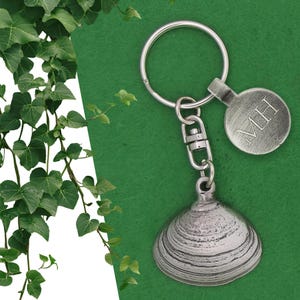 May include: A silver-coloured keychain with a clam shell pendant and a round tag bearing the initials "MH". The keychain includes a split ring and a swivel clasp. The background is a solid green colour with ivy.