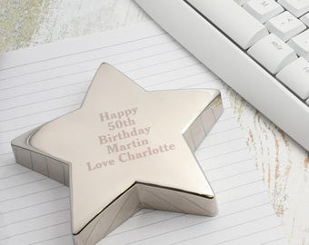 May include: A silver star-shaped desk accessory with the engraved message "Happy 50th Birthday Martin Love Charlotte." The star is on lined paper next to a white computer keyboard. The star is a decorative item.
