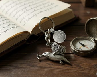 Puede incluir: Un llavero plateado con forma de colibrí con una etiqueta inicial y una brújula sobre una superficie de madera. Un libro abierto con texto manuscrito está en el fondo. El llavero presenta un diseño detallado de pájaro.