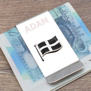 May include: A silver money clip with the name "ADAM" engraved on it. A black and white flag with a white cross is attached to the clip.