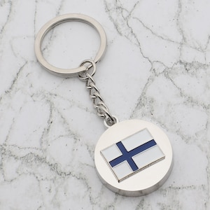 May include: A silver keychain with a round charm featuring the Finnish flag. The flag is white with a blue cross.