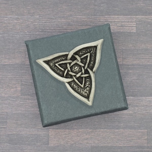 Könnte beinhalten: Ein silberfarbenes keltisches Knotenmuster auf einer teal-farbenen Box. Der Knoten ist ein dreiteiliges Symbol mit verschlungenen Linien. Die Box hat eine strukturierte Oberfläche. Der Hintergrund ist eine Holzoberfläche.