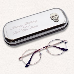 Könnte beinhalten: Ein silbernes Brillenetui mit Totenkopf-Emblem und dem eingravierten Text "Happy Birthday Jamie Love You Loads". Daneben eine runde, goldgerahmte Brille mit dunkelvioletten Bügeln.