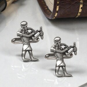 Könnte beinhalten: Zwei silberfarbene Manschettenknöpfe mit einer detaillierten Figur, die einen Bogen und einen Pfeil hält. Die Manschettenknöpfe haben einen T-Verschluss. Die Figuren sind in einer stehenden Pose, mit aufwendigen Details an Kleidung und Gesicht.