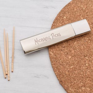 Puede incluir: Un porta palillos de dientes de plata con la inscripción "Michaels Picks" sobre un posavasos de corcho.