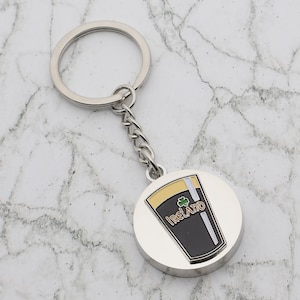 May include: Silver keychain with a round metal charm featuring a black and gold pint glass with the word "IRELAND" and a shamrock.