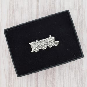 Peut inclure: Épingle à nourrice argentée en forme de train, présentée dans un coffret noir avec un intérieur en velours. L'épingle représente une locomotive à vapeur détaillée. Le coffret est posé sur une surface en bois clair.