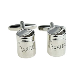 May include: A pair of silver cufflinks shaped like a can of baked beans. The cufflinks have the words "BAKED" and "BEANS" engraved on them.