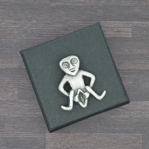 Może przedstawiać: Srebrny metalowy pin w kształcie stylizowanej postaci z dużymi oczami i wyciągniętymi ramionami, siedzącej na czarnym pudełku. Pin ma unikalny, abstrakcyjny design. Tło to ciemne usłojenie drewna.