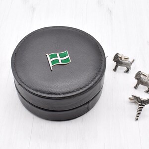 Peut inclure: Une boîte à boutons de manchette en cuir noir avec un motif de drapeau vert et blanc sur le couvercle. La boîte contient une paire de boutons de manchette en argent avec un motif de chèvre et une paire de boutons de manchette en argent avec un motif de caducée.