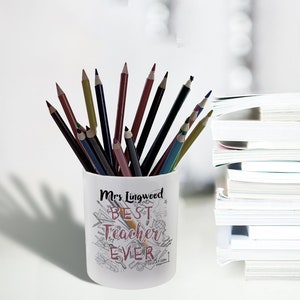 May include: A white pencil holder filled with colored pencils. The holder is decorated with the words "Mrs Lingwood BEST Teacher EVER" in a handwritten style, along with school-themed doodles. A stack of books is in the background.