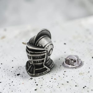 May include: A silver-coloured metal lapel pin shaped like a medieval knight's helmet with a detailed visor and crest. The pin features a metallic finish with darkened recessed areas to highlight the structural design and includes a separate metal clutch.