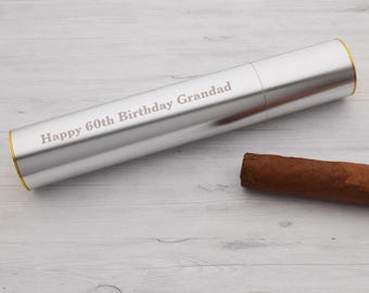 May include: A silver cigar tube with a gold trim and the inscription "Happy 60th Birthday Grandad". A brown cigar is lying next to the tube on a light wood surface.