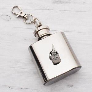 May include: A stainless steel hip flask with a silver finish, adorned with a paint bucket and brush emblem. It has a screw-on cap and a keychain clip. The rectangular flask holds approximately 148 ml. Ideal for carrying beverages, it is a practical and stylish accessory.