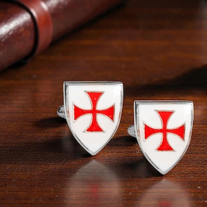 May include: A pair of silver cufflinks featuring a white shield design with a red cross emblem. The cufflinks are displayed on a dark wooden surface, with a brown leather backdrop.