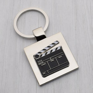 May include: Silver metal keychain with a black and white clapperboard design. The clapperboard has the words "TITLE", "DIRECTOR", "SCENE", "TAKE", "ROLL", and "DATE" printed on it.