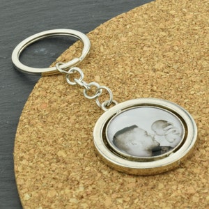 Puede incluir: Un llavero plateado con un portafotos redondo que presenta una foto en blanco y negro de un hombre y un bebé. El portafotos gira.
