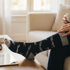 Puede incluir: Calcetines negros con talón y puntera grises, con un patrón repetido de siluetas de conejos blancos. Los calcetines tienen una banda gris en la parte superior. La persona está sentada en un sofá beige.