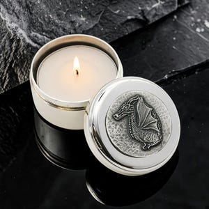 May include: A round silver-colored metal candle tin with a lit white wax candle inside. The lid is placed beside the tin and features a raised, textured pewter-toned emblem of a dragon in profile with wings folded against its scaly body.