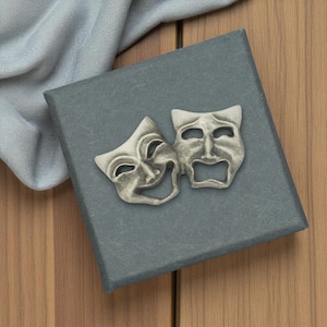 Puede incluir: Un par de máscaras de teatro de comedia y tragedia en tono plateado, sobre una caja gris. Las máscaras están detalladas con rasgos faciales y están colocadas una al lado de la otra. Un tejido azul claro está drapeado en el fondo, sobre una superficie de madera.