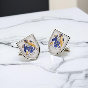 May include: Two silver cufflinks with a white shield design. Each shield displays a blue and gold crest, with the name "Holroyd" on a banner. The cufflinks are presented on a marble surface.