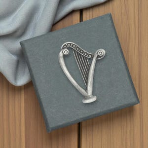 May include: A silver-colored harp pin rests on a dark gray box. The harp pin is detailed with strings and decorative elements. The box is on a wooden surface, with a light blue fabric draped nearby. The pin is a symbol of Irish heritage.