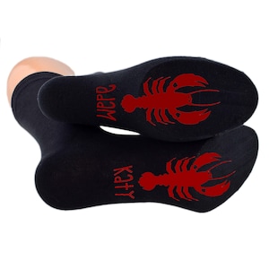 Op de afbeelding: Een paar zwarte sokken met rode kreeftontwerpen en de woorden "Katy" en "Wippo" gedrukt op de zolen.