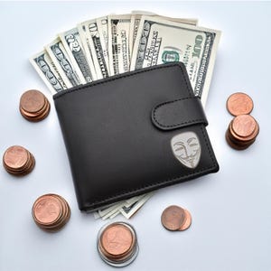 Könnte beinhalten: Eine schwarze Ledergeldbörse mit einem silbernen Metallemblem der Anonymous-Maske. Die Geldbörse ist geöffnet und mit US-Dollar-Scheinen gefüllt. Um die Geldbörse herum liegen mehrere Stapel Kupfermünzen.