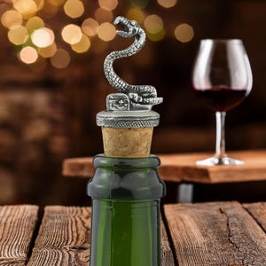May include: A silver-coloured metal bottle stopper featuring a coiled snake sculpture atop a small rectangular base. The stopper has a natural cork plug inserted into the neck of a dark green glass bottle, with a textured metal rim below the snake.