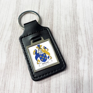 Może przedstawiać: Czarny skórzany brelok do kluczy ze srebrnym kółkiem. Prostokątna metalowa płytka przedstawia herb rodzinny z niebieskimi, żółtymi i białymi detalami oraz nazwą "Holroyd" na banerze.