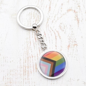 May include: A silver keyring with a round charm featuring a colourful pride flag design. The charm has a silver border and a clear protective coating.