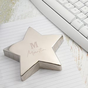 Könnte beinhalten: Ein silbernes sternförmiges Schreibtischaccessoire mit dem Buchstaben "M" und dem Namen "Martin" auf der Oberseite eingraviert. Der Stern liegt auf linierten Papier neben einer weißen Tastatur.