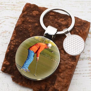 May include: A silver keychain with a round glass charm featuring a photo of a golfer putting on a green. The charm is attached to a silver key ring with a silver golf ball charm.