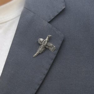 May include: A silver-toned metal brooch shaped like a pheasant in flight, featuring detailed engraved feathers and a textured finish. The bird is depicted with wings spread and a long tail, pinned securely onto the lapel of a dark gray fabric blazer.